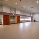 Large empty room with a row of wooden cabinets and lockers along the back wall, water fountain, and a wall clock above the cabinets; beige floor and drop ceiling.