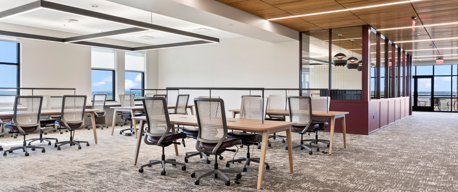 Open-plan office with rows of wooden tables and white mesh chairs under a wooden slat ceiling and large windows showing a blue sky.