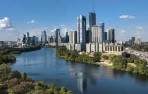 Modern city skyline with tall glass skyscrapers along a calm river and green riverbanks under a clear blue sky.