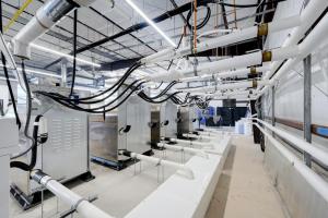 Industrial equipment room with stainless steel cabinets, numerous black cables, and overhead white insulated pipes.